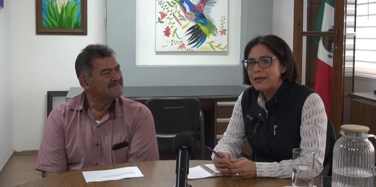Entrevista a la delegada de Bienestar en Baja California Sur, Yanssén Wiechselbaum, durante una reunión en oficina junto a un entrevistador, con bandera de México al fondo.