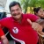 Francisco Díaz Zuñiga. Hombre adulto sonriente con camiseta roja haciendo señal de pulgar arriba, sentado en una silla blanca durante un evento al aire libre, con otras personas y vegetación al fondo.